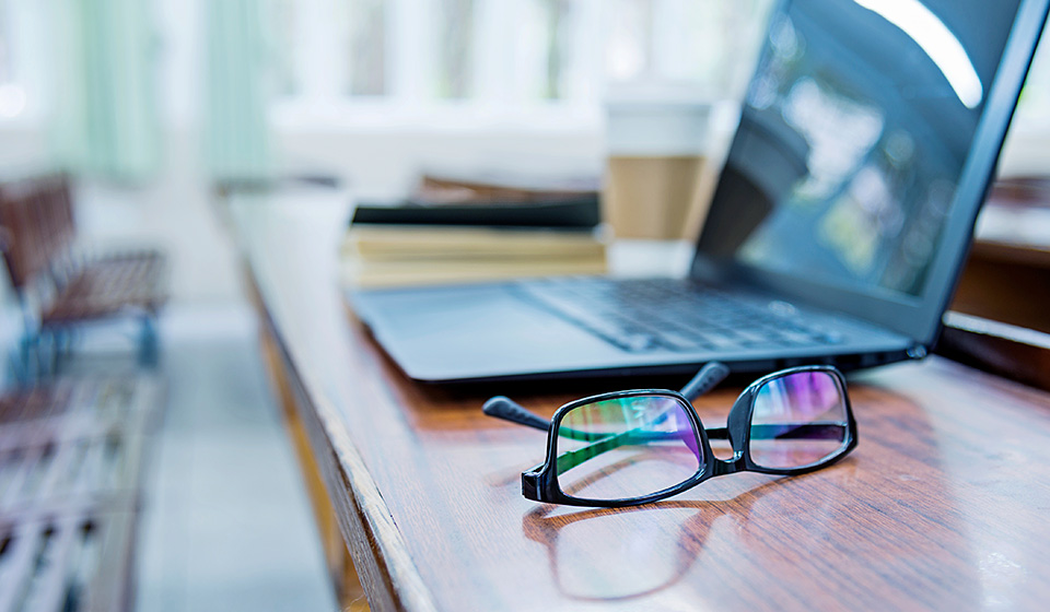 close up of an open laptop sitting on a desk with a pair of black rimmed reading glasses in the foreground