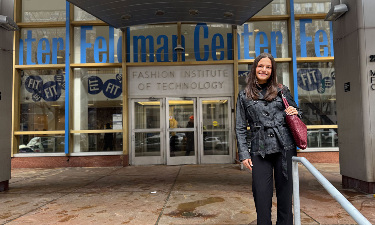 Louise King wearing a black denim coat, black pants and a red bag. She has long dark hair and is smiling. She is standing in from of the Fashion Institute of Technology in New York City.