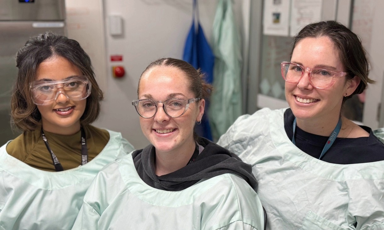 Three female researchers in lab coats smiling at the camera.
