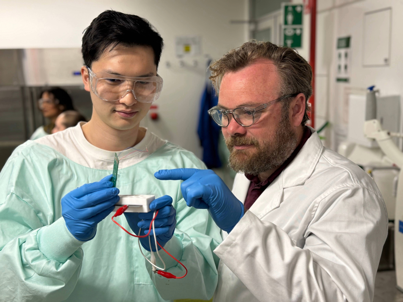 Two researchers in lab coats and gloves examining a small white device with wires attached.
