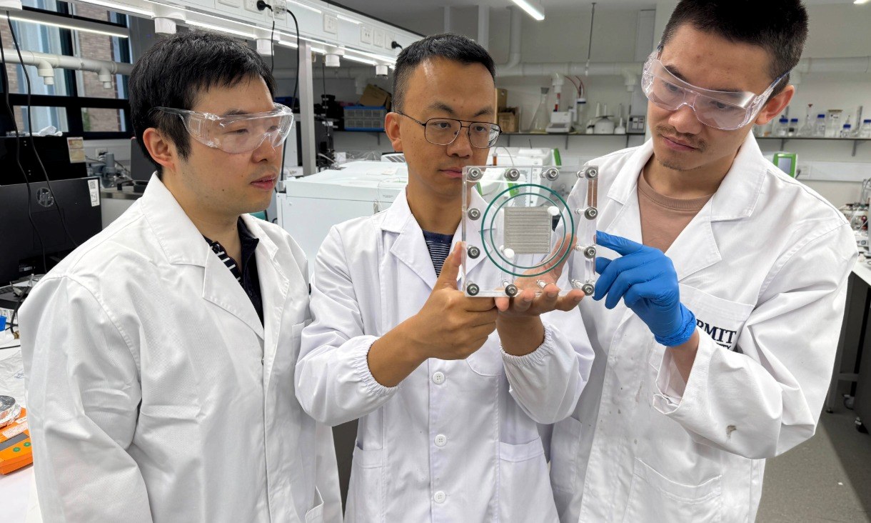 Three researchers in white lab coats standing in an RMIT laboratory. The person in the centre is holding a transparent, square device with metal bolts and a circular green seal. Laboratory equipment and benches are visible in the background.