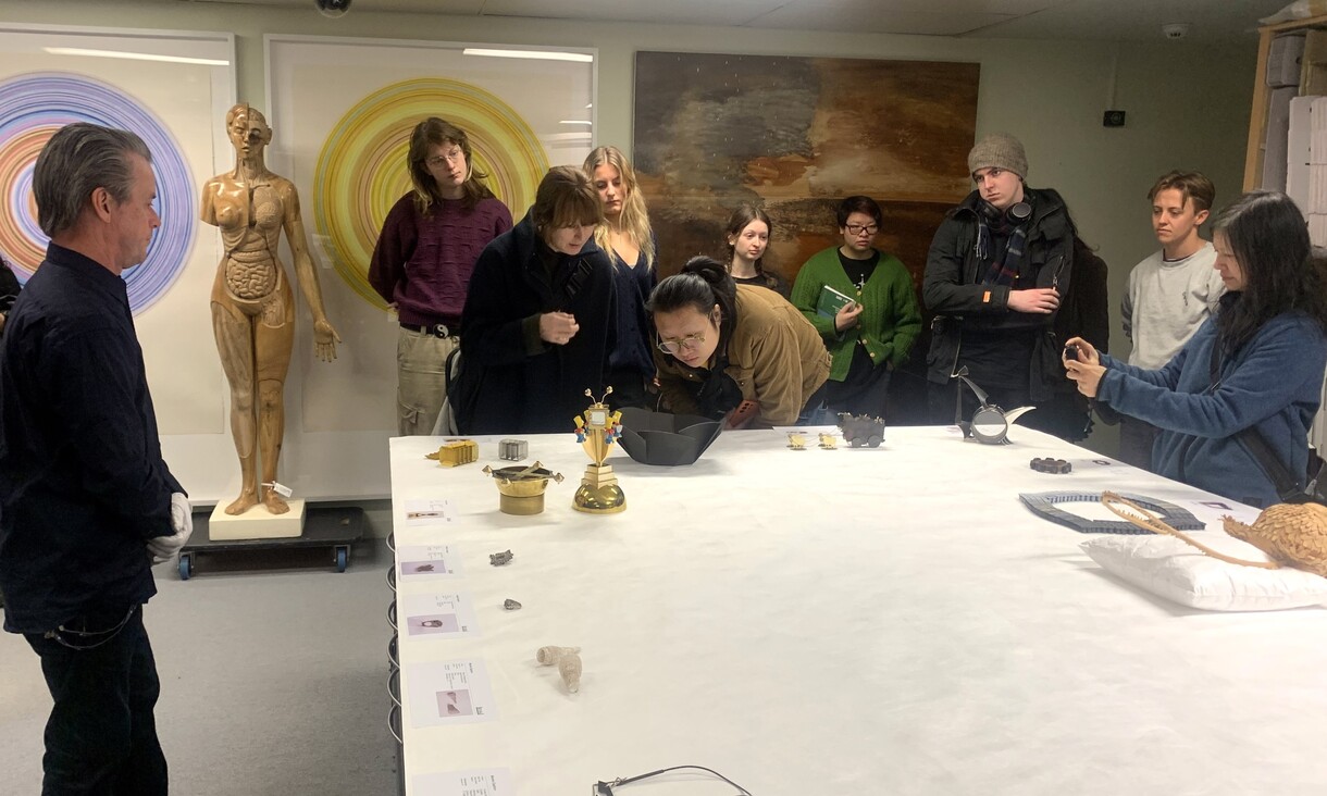 A group of ten people stand in a gallery, looking at items on a table. 