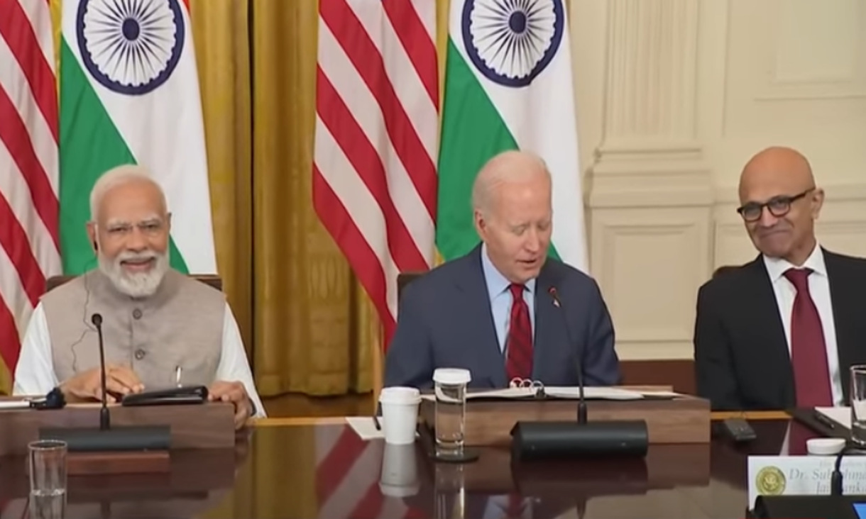 US President Biden sitting at table flanked by Indian Prime Minister Narendra Modi and  Microsoft CEO Satya Nadella with Indian and US flags in the background