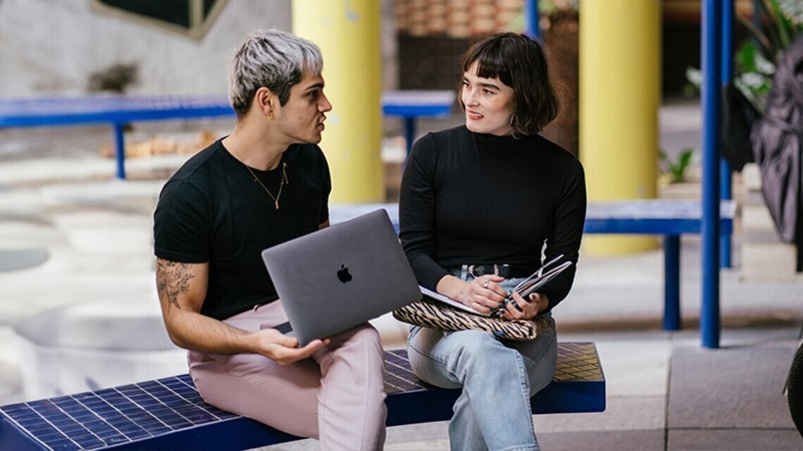 A blonde man with a laptop on his lap sitting beside a dark-haired, smiling woman with a fringe who is holding a notebook. They sit on a bright blue bench in an open outdoor area. 