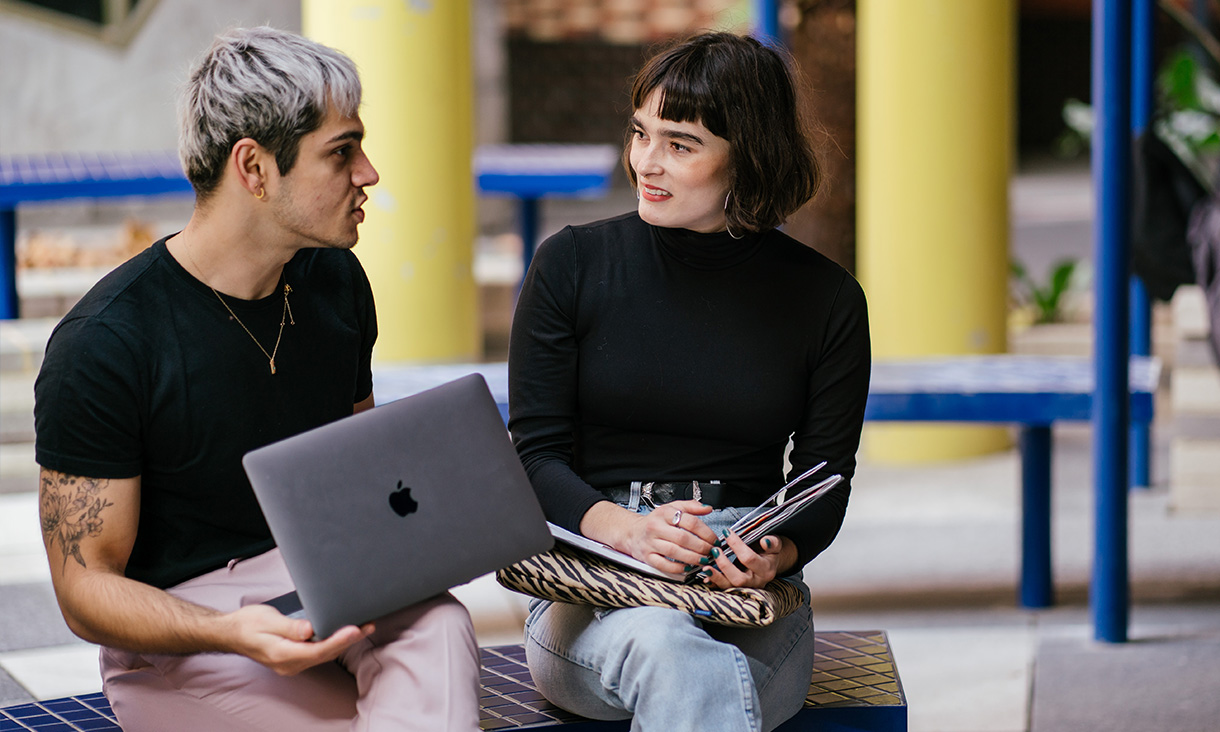 A blonde man with a laptop on his lap sitting beside a dark-haired, smiling woman with a fringe who is holding a notebook. They sit on a bright blue bench in an open outdoor area. 