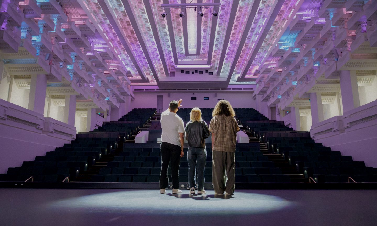 Three people stand on stage at the capitol, looking out at the empty seats