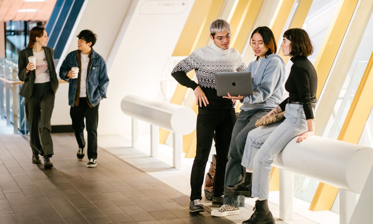 Three students standing in a hallway, one is holding a laptop the other two are looking at the screen. They are discussing what they are seeing. 