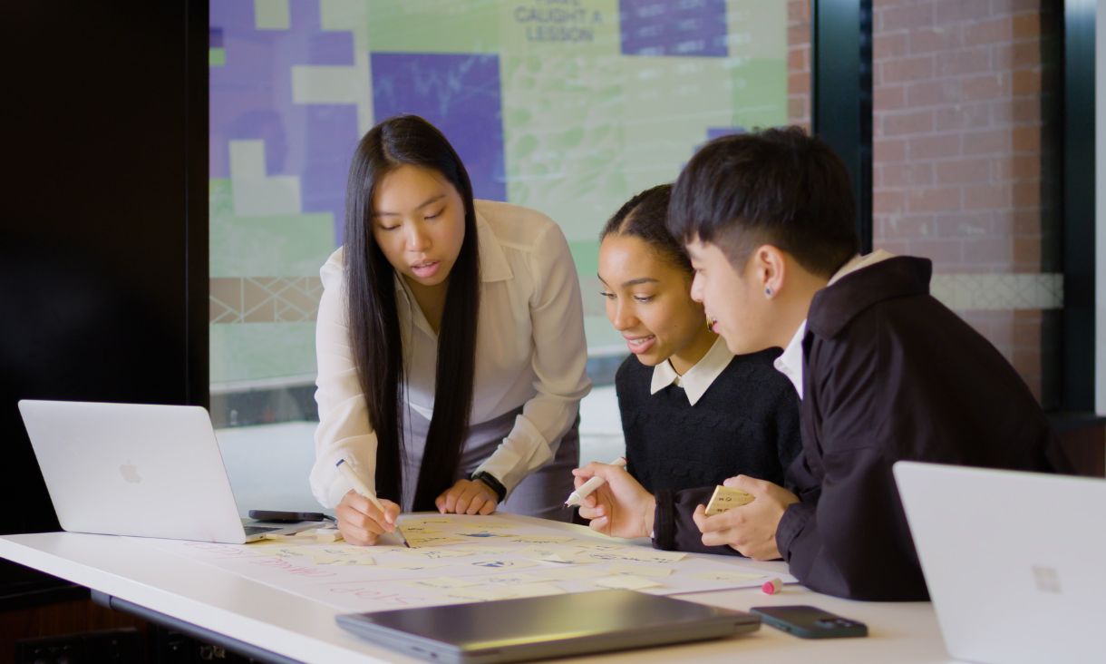 two students sit at a table looking at chart spread out on the table. Another student leans over the chart making notes wit ha pen. They are dressed in smart casual clothes. Laptops are out on the table as well. 