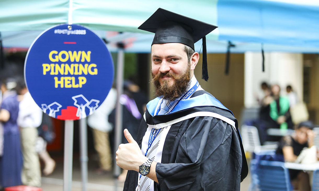 Male graduate smiles and camera and gives a thumbs up in frony of a sign that reads gown pinning help.