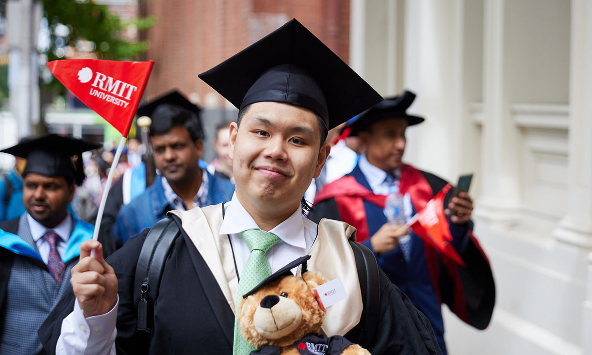 Male graduate in academic dress smiles at the camea and waves an RMIT flag