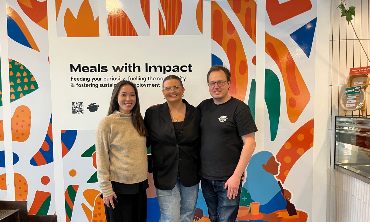 Three people pose for a photo in front of a sign which reads "Meals with Impact"