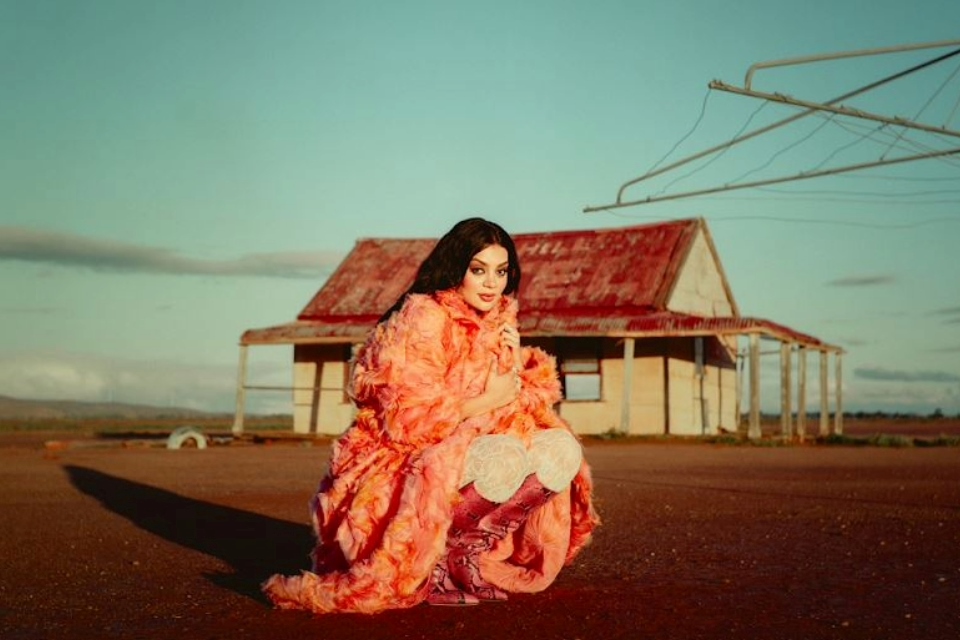 Woman dressed in bright orange colors kneeling in front of an old rustic home.