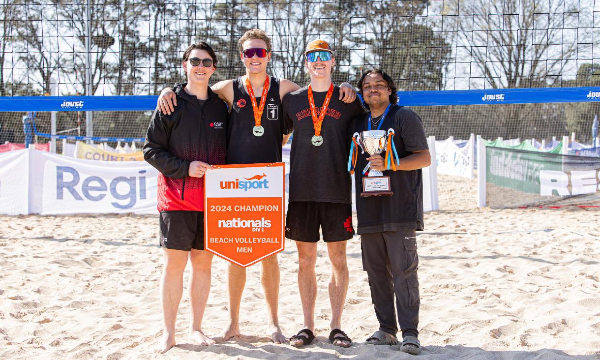 Four volley ball players pose on the sand next to the nest with trophies and medals.