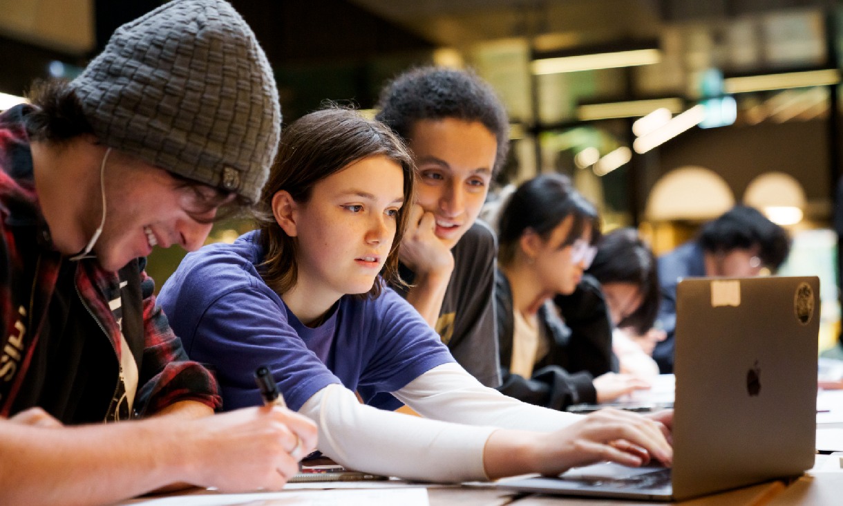 Line of students seated along a table, focused on writing and reading from a laptop