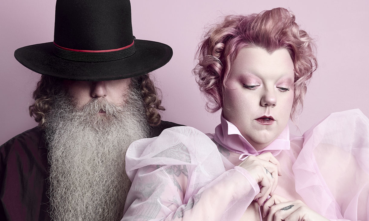 A man stands on the left, he has a long grey beard and shoulder length brown hair. He wears a black bowler hat with a red ribbon that covers his eyes and a maroon shirt. A woman stands in front to the right, she looks away from the camera and wears all pink, including her makeup and curly hair. 