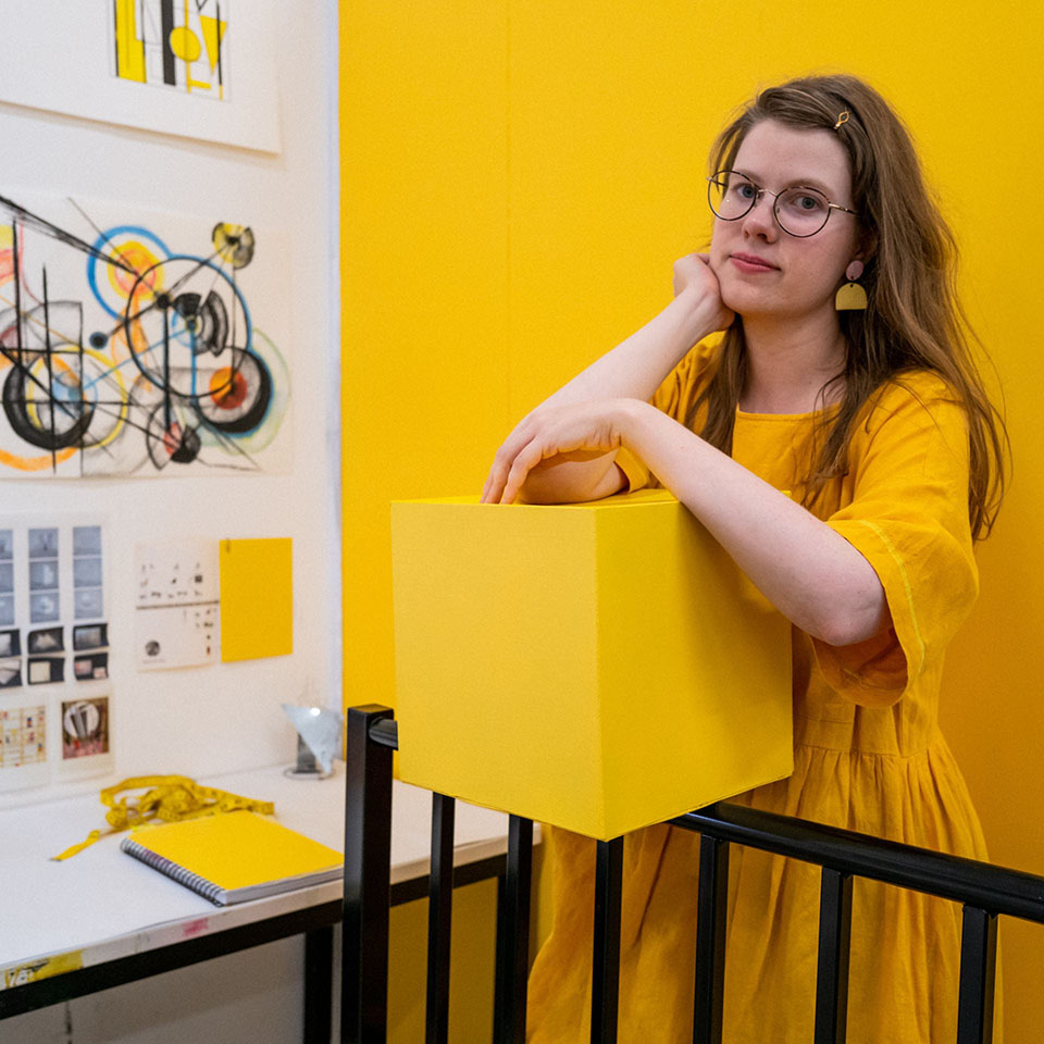 A person in yellow leans on a yellow box in a fine arts studio.