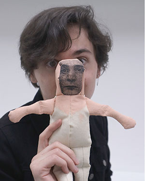 A person holding a cloth doll with a printed face in front of their own face.