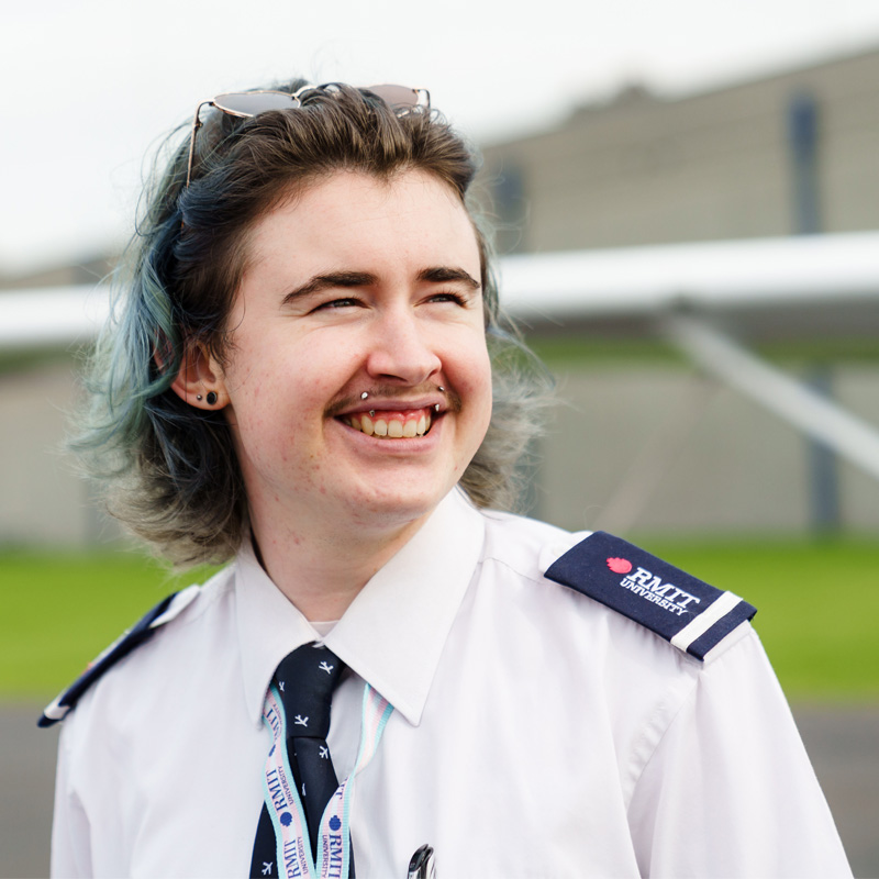 Portrait of Otto smiling at the camera. He wears a pilot uniform and has shoulder length brown hair with blue highlights.