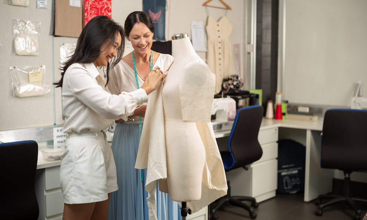 A female lecturer helps a female fashion student pin fabric to a mannequin in a fashion lab.