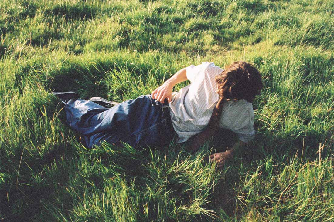 A person in jeans, a white short-sleeved shirt and a brown tie lounges in long green grass.