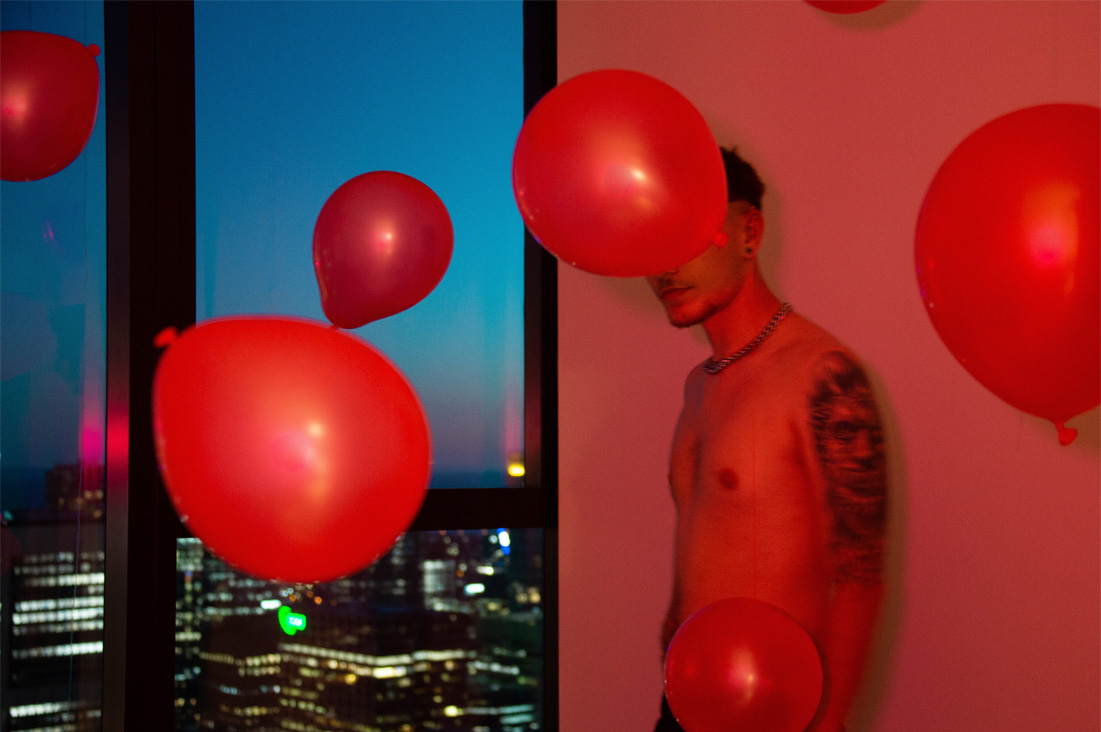 A red light shines on a shirtless man leaning against a wall next to a window. Red balloons fall around him, one covers his face. Outside the window is early evening and a view of a cityscape.