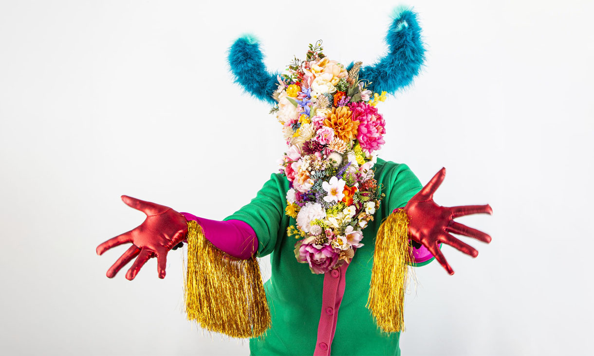 A person with their head and chest covered by a floral arrangement. They have blue fluffy antlers and are wearing a green and pink onesie with red gloves. Yellow tassels are attached to their arms.