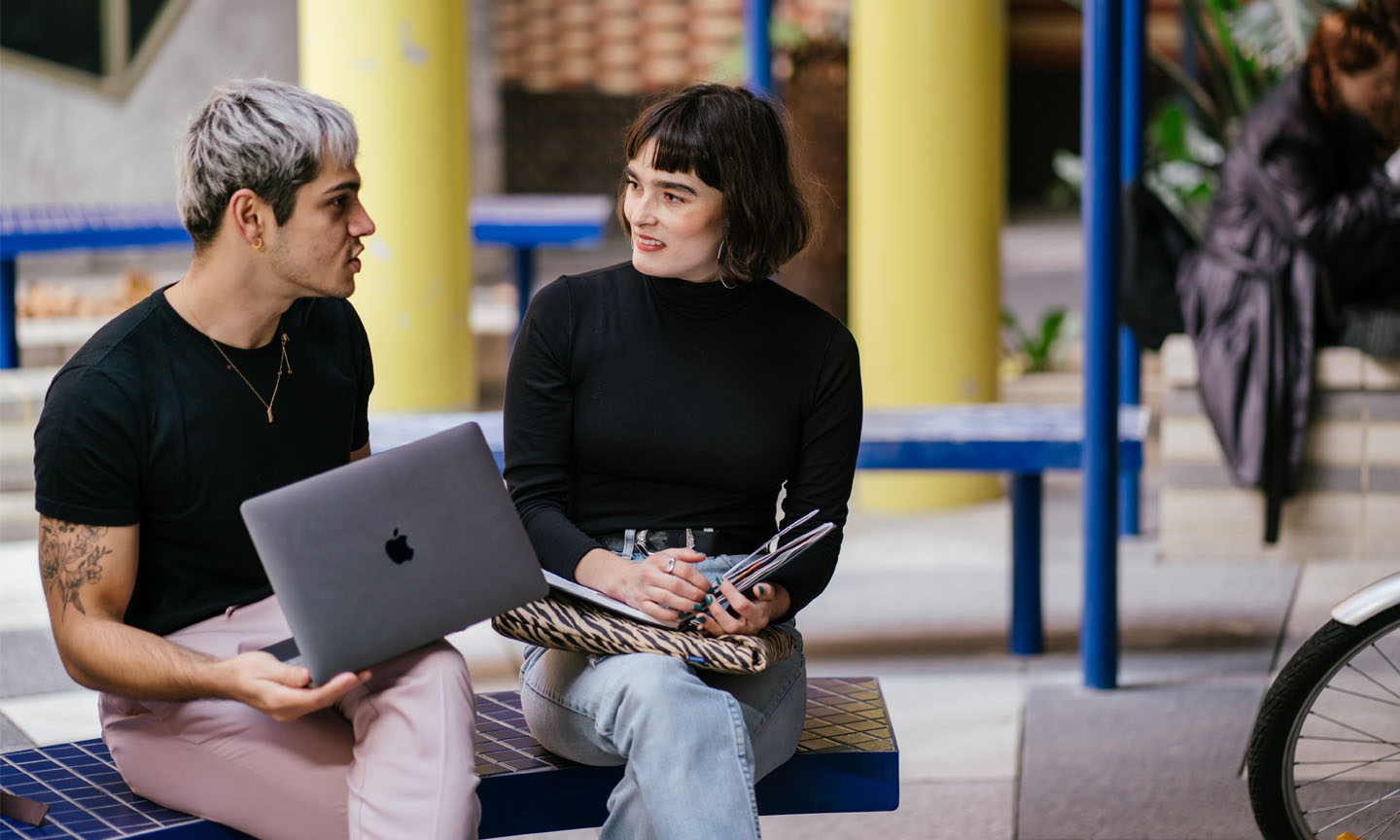 Two RMIT students sitting on a bench discussing their notes. One student has a notebook while the other has a laptop