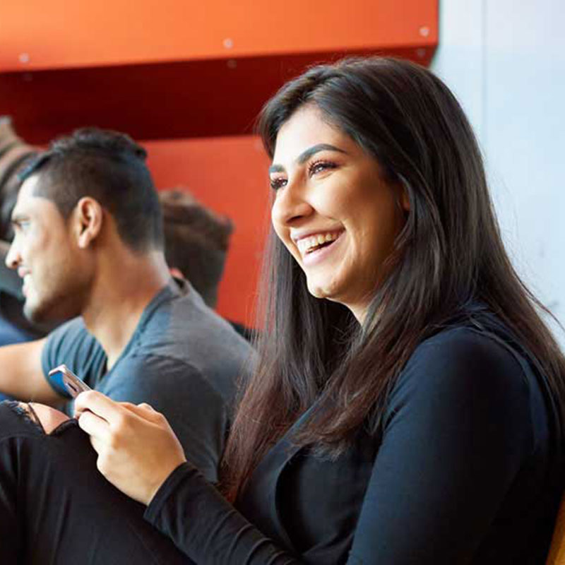 A female student smiling and holding a mobile phone in front of a red background with male student.