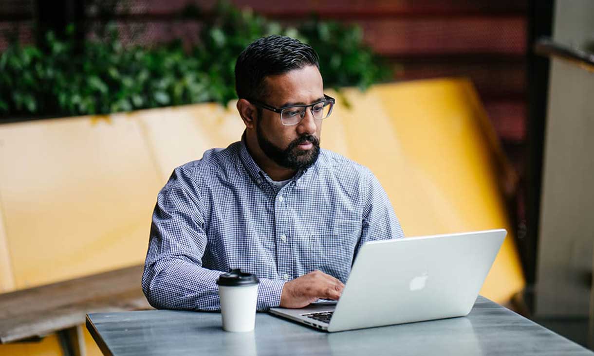 Student sitting at table with laptop and coffee in front of yellow background.