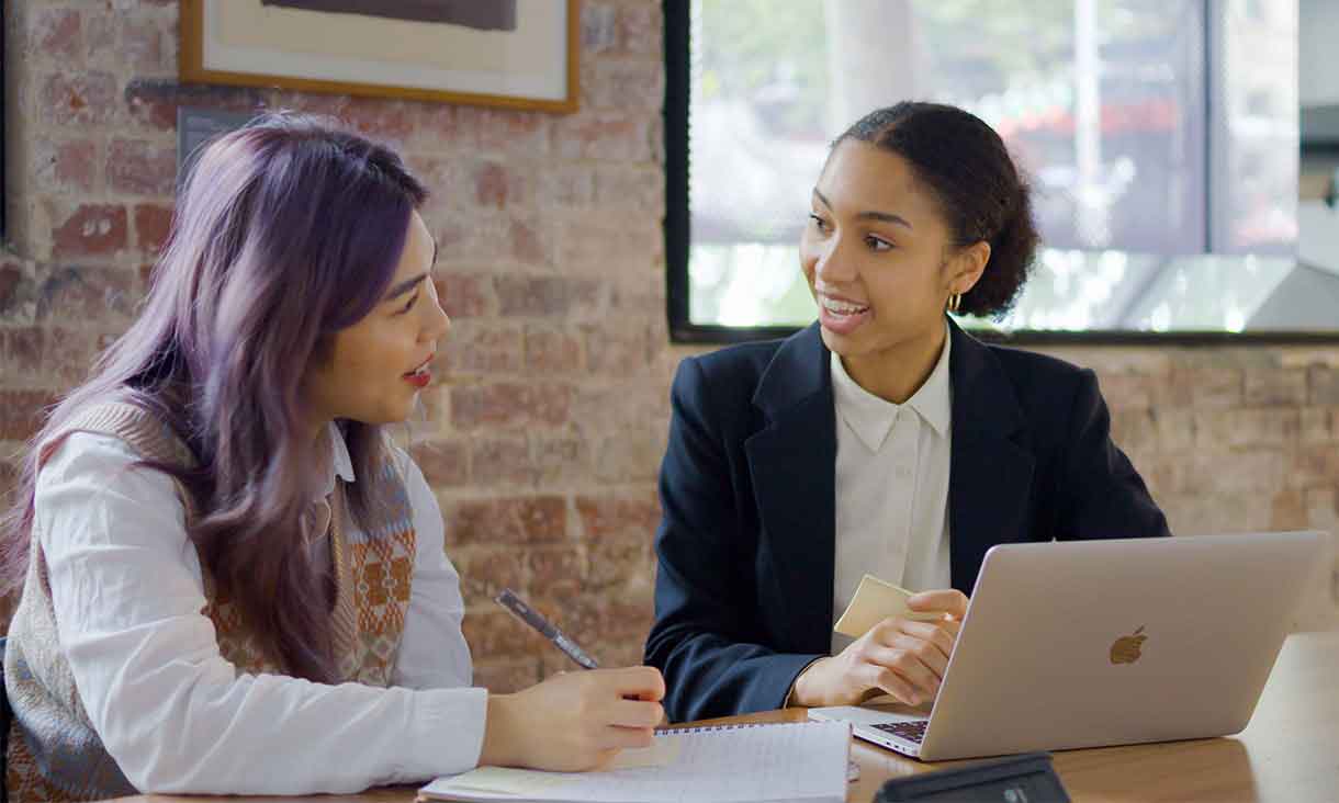 Two female students sitting at a wooden table with laptop, pen and notebook.