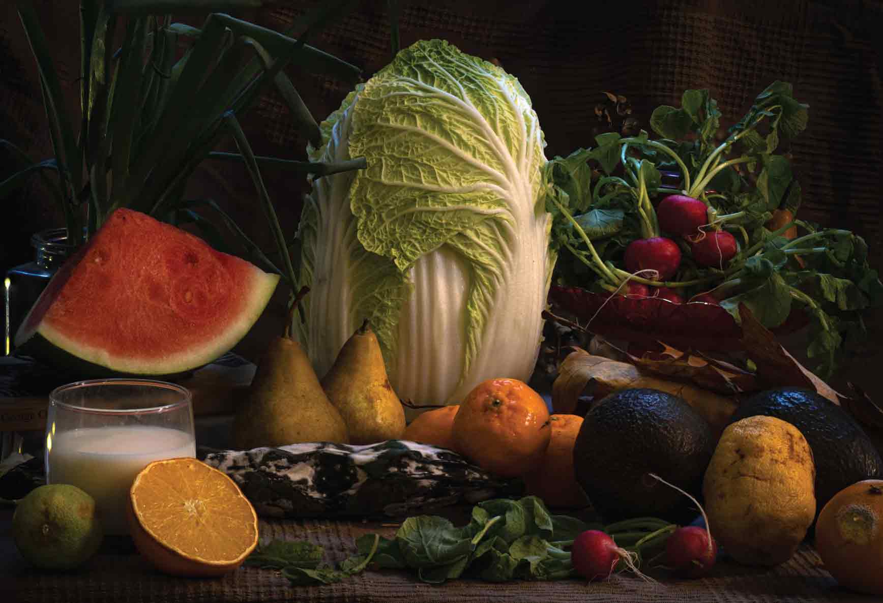 a still life photograph with soft dramatic lighting depicting vegetables, fruit, and a glass of milk arranged on a table.