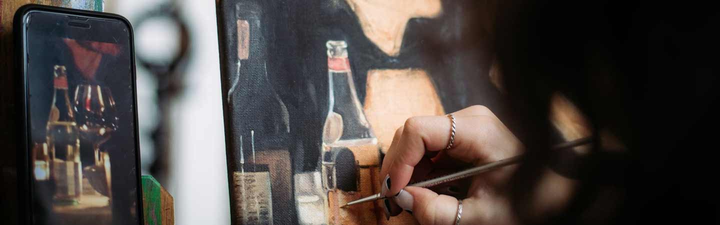 a hand holding a fine brush painting a bottle. A photograph of the bottle is displayed on a mobile device resting against an easel.