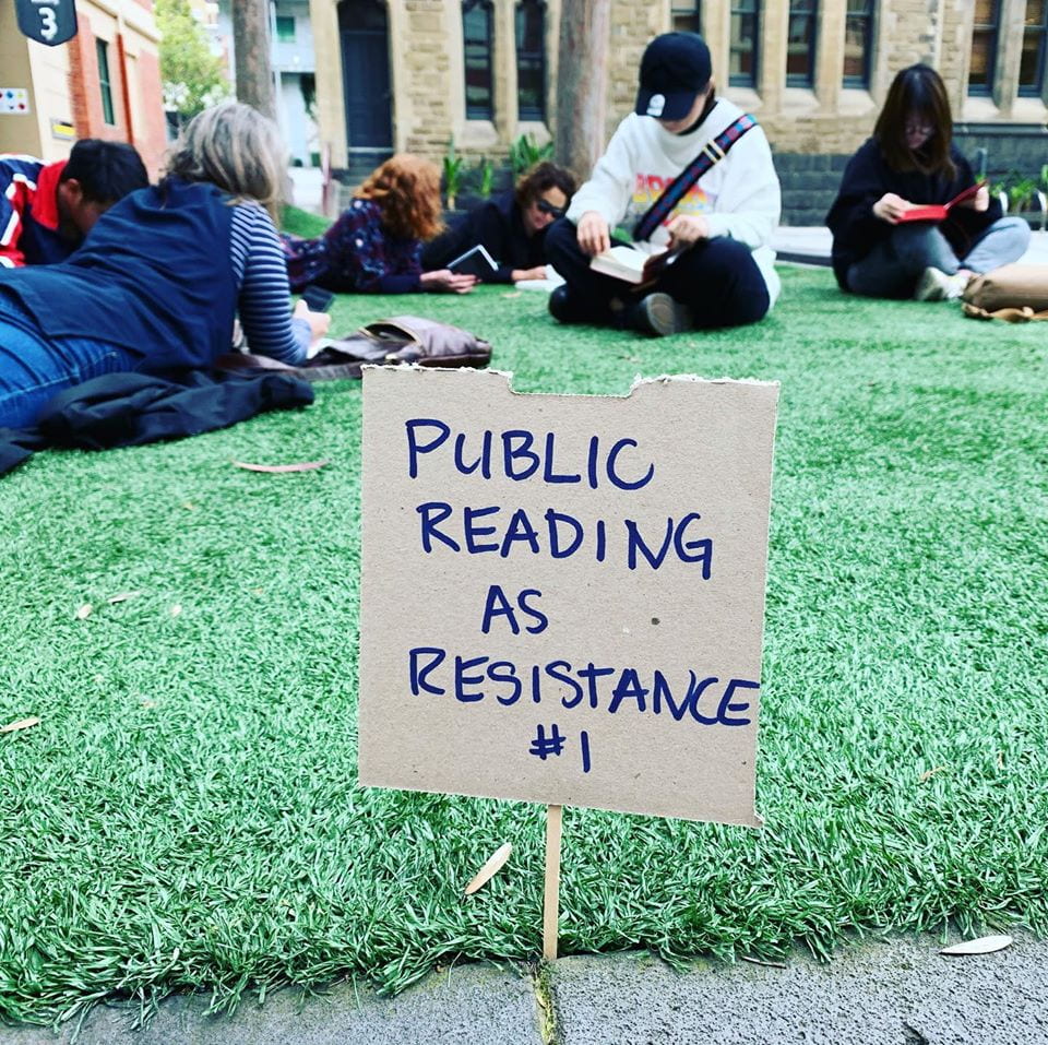 a photograph of people sitting outside on grass reading books. At the front of the image is a small handwritten sign on cardboard that says, 'PUBLIC READING AS RESISTANACE #1'.