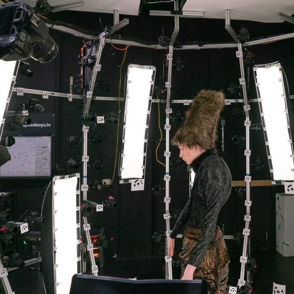 a figure wearing a black short, brown skirt and tall brown fluffy hat stands within a photogrammetry rig consisting of bars and cameras.