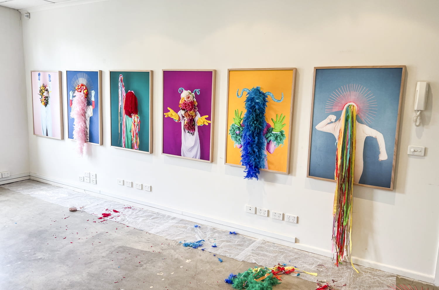 six framed artworks hanging on a wall. Each artwork depicts a figure wearing different colourful costume that includes a headdress and covers the face. elements of the costume, such as feathers and fake flowers, have been attached to the surface of each artwork.