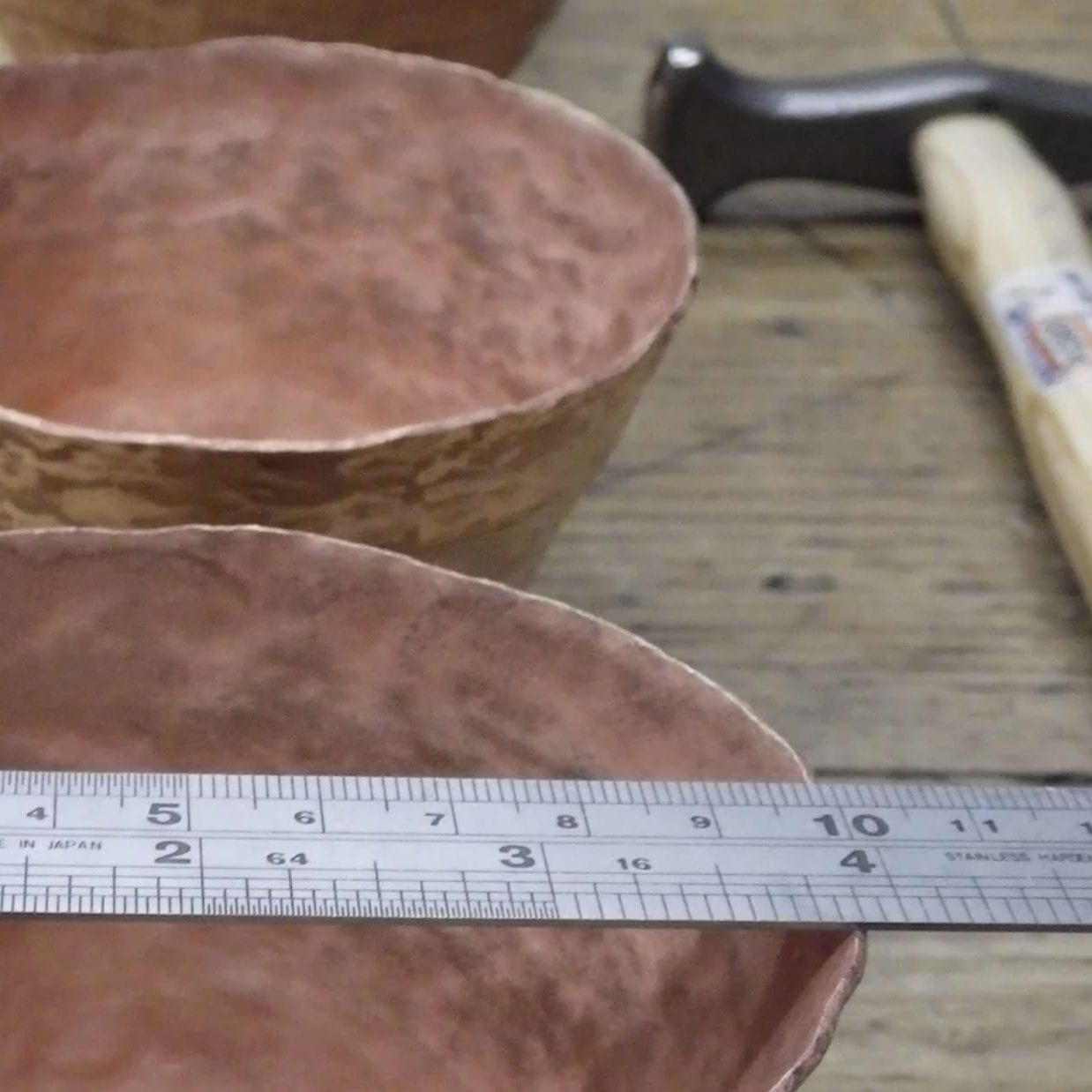 photograph depicting details of two pale pink hand-made bowls sitting on a wooden table. A hammer sits on the right-hand side. A ruler sits across the top of the closer bowl.