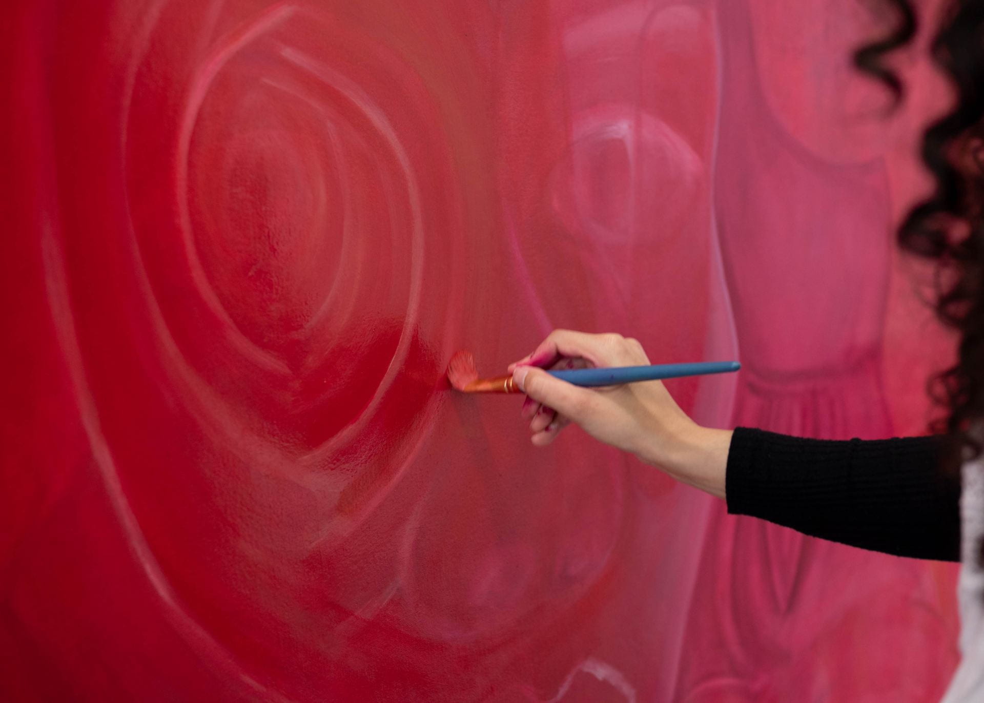  a hand holding a paintbrush reaching towards a red painting.