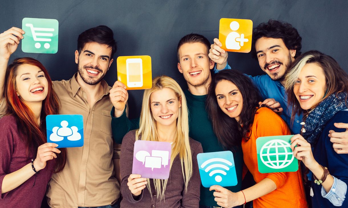 Stock image of people smiling and holding up cards with icons representing modern technology. Icons include iPhone and wifi strength symbol.