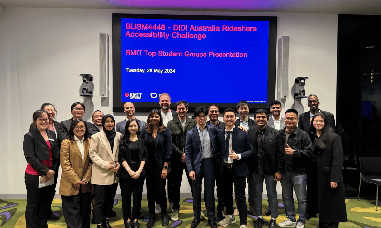 A group of students and industry professionals standing in front of a projected screen reading 'BUSM4448 - DiDi Australia Rideshare Accessibility Challenge RMIT Top Student Groups Presentation'