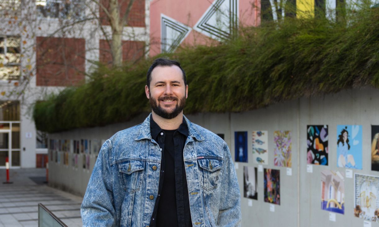 Man in black shirt and denim jacket in front of concrete wall with shrubbery.