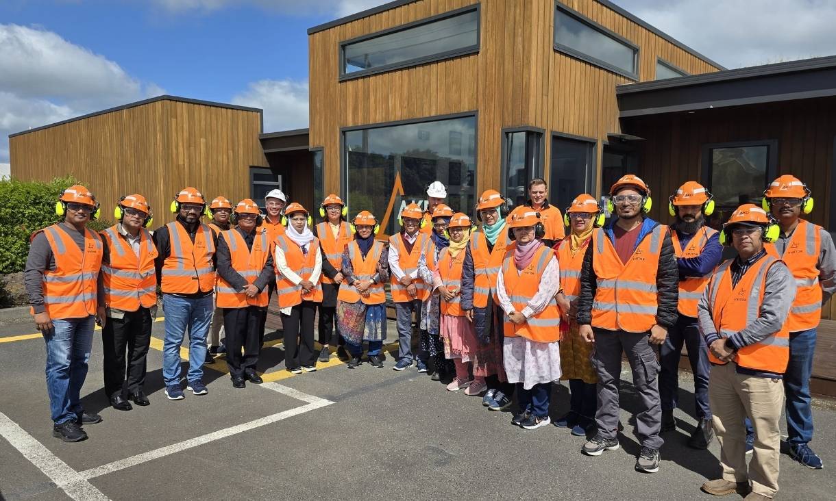 Large group of researchers in orange hi-vis vests and hard hats, posing in front of wood-panelled building.